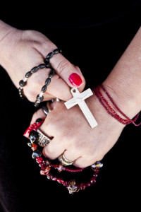 Two white-skinned hands wrapped in red prayer beads and a black rosary.