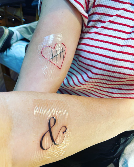 Two white-skinned arms displaying new tattoos, one of a heart and one of an ampersand.