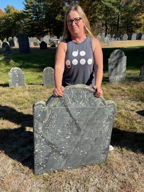 A blonde white woman standing in an old graveyard behind a tombstone with hard-to-read old text.