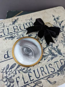 A miniature painting of a blue eye in a round gold frame, atop a vintage photo album with lettering in French.